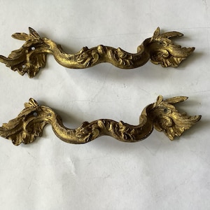 May include: Two antique gold-colored drawer pulls with ornate, leaf-like designs. Each pull features a curved handle with detailed floral and foliate embellishments. The hardware is likely made of metal and shows signs of age.