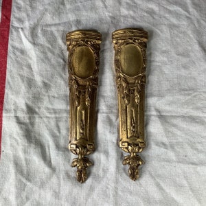 May include: Two gold-colored metal wall sconces with ornate designs. Each sconce has a rectangular shape with a raised oval in the center. The sconces have a detailed floral pattern and a decorative bottom.