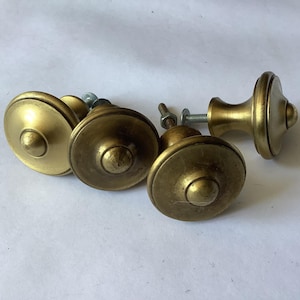 May include: A set of four circular brass cabinet knobs with a smooth, metallic finish. Each knob features a raised center dome and a flat, wide base. The knobs are equipped with threaded metal bolts and nuts extending from the back for installation.