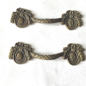 May include: Two matching brass drawer pulls featuring a curved handle with a leaf pattern. Each end of the pull has a circular medallion displaying a profile portrait of a person, topped with a decorative bow and ribbon motif in a metallic finish.