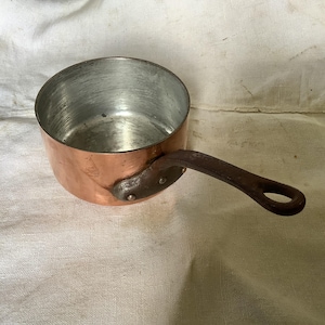 May include: A copper saucepan with a dark brown handle. The saucepan has a shiny copper finish and a small, round handle.