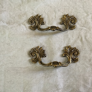 Può includere: Due tiretti per cassetti in ottone decorati con un design a rotolo. I tiretti hanno uno stile classico e vintage.