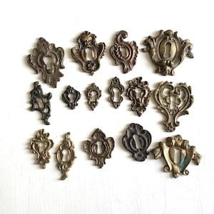 May include: A collection of antique brass keyhole covers in various ornate designs. The covers feature intricate floral and scrollwork patterns, with a range of sizes and finishes, from dark bronze to aged gold. These decorative hardware pieces are ideal for vintage furniture restoration.