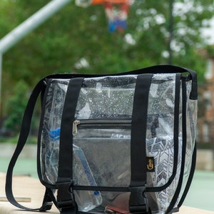 Puede incluir: Bolso transparente de pl&aacute;stico con correa cruzada, con correas y hebillas negras. El bolso tiene un bolsillo con cremallera y est&aacute; lleno de varios art&iacute;culos, incluyendo una botella de agua, un tel&eacute;fono y una peque&ntilde;a bolsa.