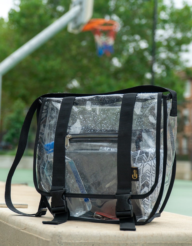 Puede incluir: Bolso transparente de pl&aacute;stico con correa cruzada, con correas y hebillas negras. El bolso tiene un bolsillo con cremallera y est&aacute; lleno de varios art&iacute;culos, incluyendo una botella de agua, un tel&eacute;fono y una peque&ntilde;a bolsa.