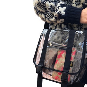 Puede incluir: Un bolso transparente de pl&aacute;stico con correa cruzada, con correas y hebillas negras. El bolso tiene un bolsillo con cremallera en la parte delantera y est&aacute; lleno de varios art&iacute;culos, incluyendo una bolsa estampada en rosa y blanco.