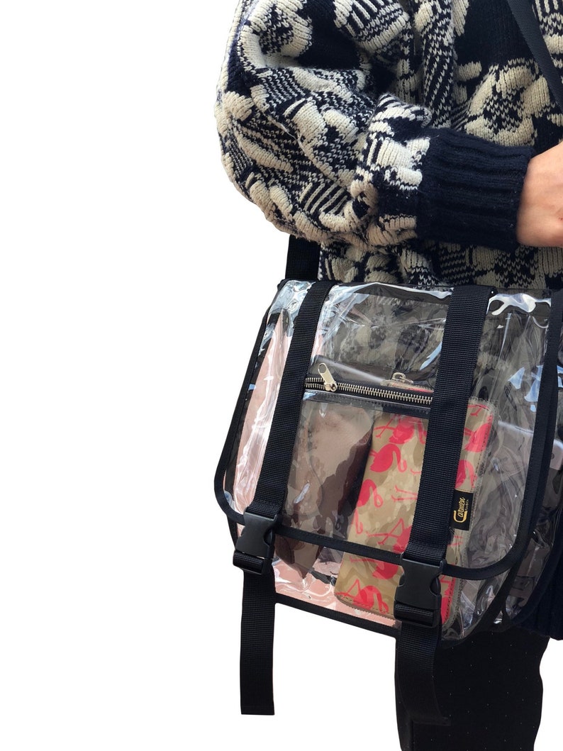 Puede incluir: Un bolso transparente de pl&aacute;stico con correa cruzada, con correas y hebillas negras. El bolso tiene un bolsillo con cremallera en la parte delantera y est&aacute; lleno de varios art&iacute;culos, incluyendo una bolsa estampada en rosa y blanco.