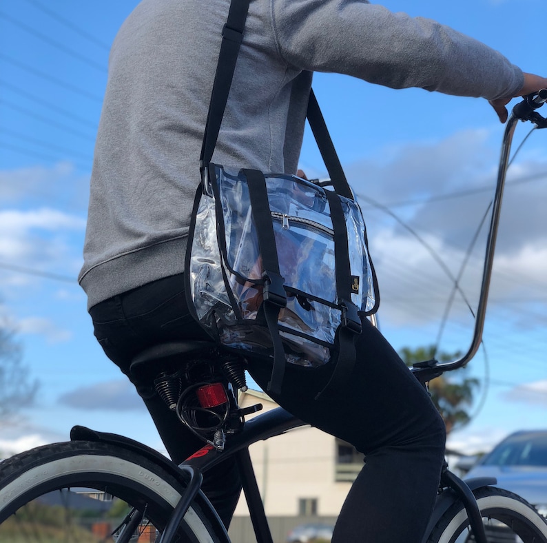 Puede incluir: Una persona montando en bicicleta con una bolsa de mensajero de pl&aacute;stico transparente con correas negras. La bolsa se lleva sobre el hombro y se asegura con una hebilla.