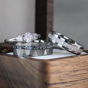 Peut inclure: Trois bagues argentées présentées dans une boîte en bois. Une bague comporte une pierre ronde centrale avec deux pierres latérales plus petites. Une autre bague a un anneau facetté avec une incrustation sombre et texturée. La troisième est un anneau serti de petites pierres claires.