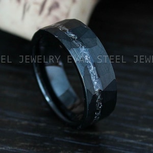 May include: A black ring with a faceted surface and a central inlay of shimmering, textured material. The band features a dark, matte finish with a jagged, crystalline-like stripe running horizontally around the centre. Text reads EVAN STEEL JEWELRY.