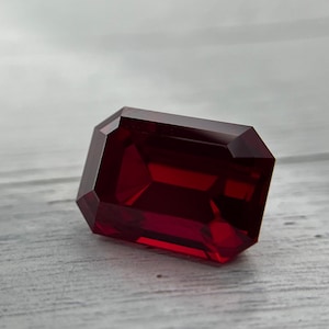 May include: A faceted, rectangular-cut, deep red gemstone. The gem has multiple facets that reflect light, creating a brilliant shine. The stone is set against a light gray and white background.
