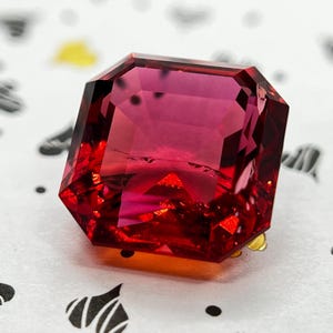 May include: A faceted, square-cut gemstone with a deep, vibrant red and magenta hue. The stone features sharp, geometric edges and a highly reflective, transparent surface that displays internal light refraction and clear, precise cut facets.