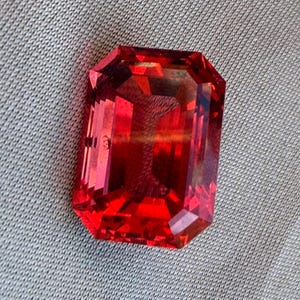 May include: A vibrant red gemstone with an emerald cut, featuring a rectangular shape and faceted surfaces that reflect light. The stone displays deep crimson and orange hues throughout its transparent body, showcasing precise geometric edges.