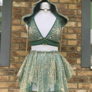 Puede incluir: Un conjunto verde de dos piezas con una parte superior con capucha y una falda, exhibido en un maniquí. La parte superior presenta diseños dorados de estrellas y celestiales, con una falda a juego. Una cadena y un collarín dorados completan el look. El conjunto está hecho de tela suave y fluida.