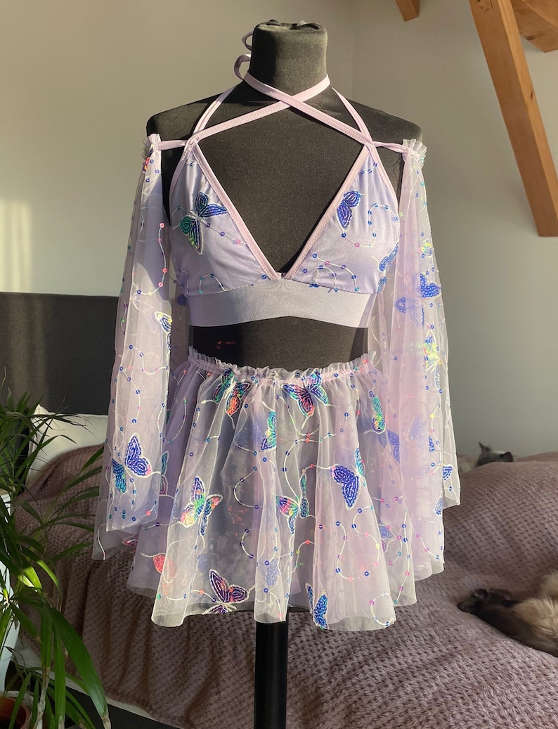 May include: A lavender-colored mesh top and skirt set with butterfly embroidery. The top has a halter neckline and long sleeves. The skirt is a mini skirt with a ruffled hem.