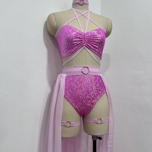 May include: A two-piece outfit featuring a shimmering pink metallic crop top with a pentagram-style strap design and a matching high-waisted bottom. The set includes a choker, a belt with a silver ring, and two leg garters, all accented by a sheer light pink skirt.