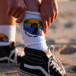 May include: White socks with a graphic of a bat surfing and the text "Beach Bats + Beer". The socks are being pulled up on a person's leg, and the person is wearing black high-top trainers.