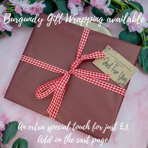 May include: A square gift box wrapped in solid burgundy paper, tied with a red and white gingham ribbon bow. A small rectangular kraft paper tag is attached, featuring the printed text A LITTLE SOMETHING SPECIAL Just For You with floral accents.