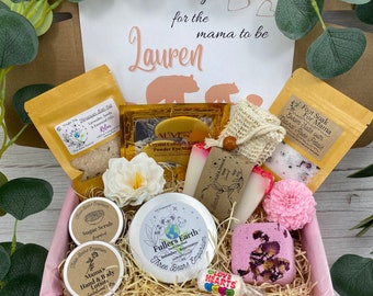 Puede incluir: Una caja de regalo para una futura mamá llamada Lauren que contiene varios artículos de cuidado personal, incluyendo sales de baño, una máscara de ojos dorada, un exfoliante de azúcar, loción para manos y cuerpo, una mascarilla de arcilla, jabón y una bomba de baño rosa.