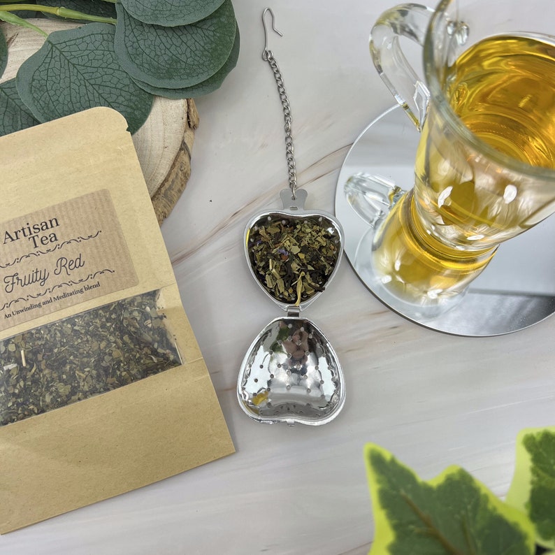 May include: A stainless steel heart-shaped tea infuser with a chain. The infuser is filled with loose leaf tea and is sitting on a white surface. The tea is labeled "Artisan Tea Fruity Red An Unwinding and Meditating Blend".