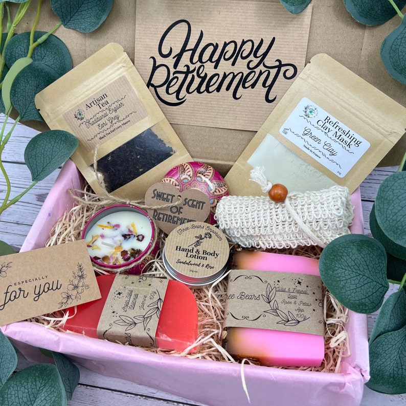 May include: A pink gift box filled with various self-care items, including a green clay face mask, a tea bag, a candle, a hand and body lotion, a bar of soap, and a small bag of tea. The box is decorated with a tag that says "Happy Retirement".