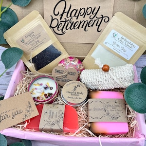 May include: A pink gift box filled with various items for a retirement gift. The box contains a candle, soap, lotion, tea, a clay mask, a washcloth, and a small decorative tin. The box is decorated with a tag that says "Happy Retirement".