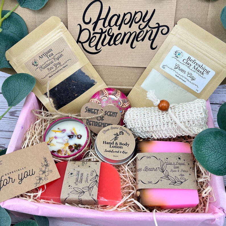 May include: A pink gift box filled with various items for a retirement gift. The box contains a candle, soap, lotion, tea, a clay mask, a washcloth, and a small decorative tin. The box is decorated with a tag that says "Happy Retirement".