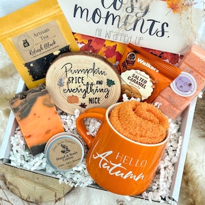 May include: A gift box containing a yellow bag of Artisan Tea, a wooden slice with the text Pumpkin Spice and everything nice, a pack of Walkers Salted Caramel biscuits, a bar of orange and black soap, lotion, and an orange mug with Hello Autumn.