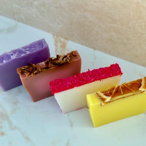 May include: Four rectangular bars of soap arranged in a row. The first is solid purple, the second is dusty rose topped with dried flower petals, the third is white with a bright pink textured top, and the fourth is yellow with a dried orange slice.