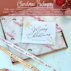 May include: A cardboard box containing white tissue paper with red and green holly and Merry Christmas text, a white greeting card with gold snowflakes and Merry Christmas and Happy New Year text, and a ribbon with Happy Christmas text.