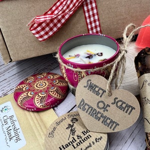 May include: A gift box filled with a variety of items, including a pink candle with a floral design, a heart-shaped tag that reads "Sweet Scent of Retirement", a small tin of hand and body lotion, and a brown paper bag with a clay mask.