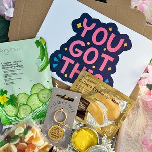 May include: A gift box filled with self-care items. Includes a cucumber facial mask, a sticker with the text "YOU GOT THIS", a gold-colored eye mask, a clear quartz keychain, and a small container of yellow balm.