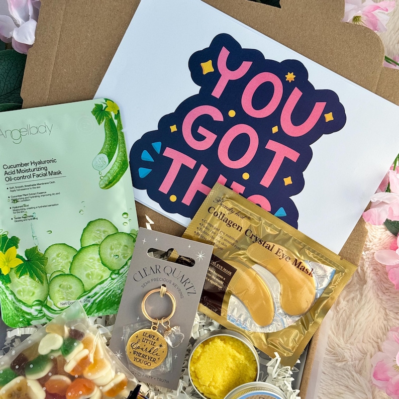 May include: A gift box filled with self-care items. Includes a cucumber facial mask, a sticker with the text "YOU GOT THIS", a gold-colored eye mask, a clear quartz keychain, and a small container of yellow balm.