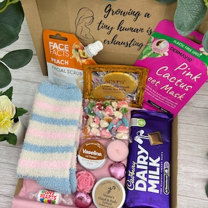 Könnte beinhalten: Eine Geschenkbox mit einem Face Facts Pfirsich-Gesichtspeeling, einer DermaV10 Pink Cactus Tuchmaske, Aliver Gold-Kollagen-Augenpads, flauschigen blau-rosa gestreiften Socken, einer Tüte Herzbonbons, Vaseline Lippenpflege, einer Kerze und Cadbury Dairy Milk.