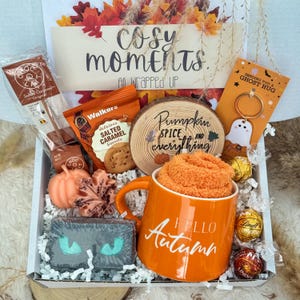 May include: A gift box containing an orange mug with Hello Autumn text, orange fuzzy socks, a wooden slice with Pumpkin Spice and everything text, Walkers salted caramel biscuits, a Choc-O-Lait stick, a ghost keychain, chocolates, and soaps.