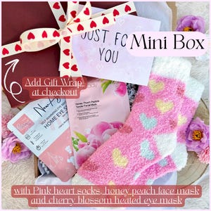 May include: A gift box containing a pair of fuzzy pink socks with yellow and blue hearts, a Honey Peach Peptide Tender Facial Mask, and a New Day Self Warming Home Eye Mask with cherry blossom scent, tied with a white ribbon with red hearts.