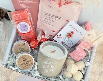 May include: A Christmas-themed gift box containing a variety of self-care products. The box includes a candle, face masks, wax melts, hand and body lotion, chocolates, and marshmallows. A Christmas card is also present.