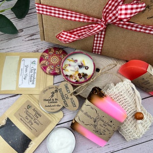 May include: A gift box filled with self-care items, including a pink and orange soap bar, a small candle with dried flowers, a green clay mask, a tea bag, a hand and body lotion, and a small round tin with a floral design.