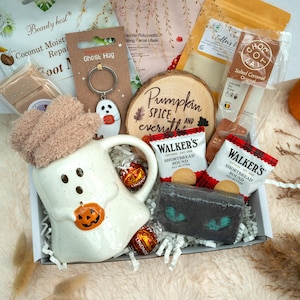 May include: A gift box containing a white ghost-shaped ceramic mug with a pumpkin, a pair of fuzzy tan socks, a ghost keychain, two Walker's shortbread packets, a dark grey bath bomb, a hot chocolate stick, and a wood slice with Pumpkin Spice text.
