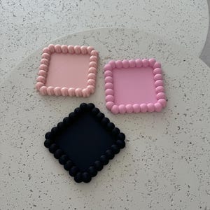 Op de afbeelding: Drie vierkante siliconen onderzetters met een parelrand. Eén onderzetter is roze, één is lichtroze en één is zwart.