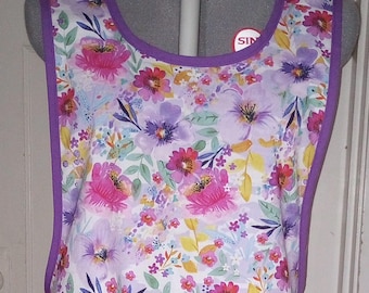 May include: A purple and white floral apron with a purple trim. The apron has a large pocket on the front.