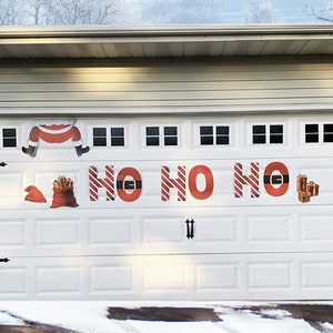 May include: A white garage door with a red and white "HO HO HO" banner and Santa Claus decorations. The decorations include a Santa hat, a bag of gifts, and a Santa Claus figure hanging upside down.