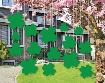 Può includere: Decorazioni a forma di trifoglio verde appese davanti a una casa con un albero in fiore rosa. I trifogli sono sospesi da fili bianchi, creando un'atmosfera festosa per il giorno di San Patrizio. La casa ha un rivestimento grigio e un prato verde.