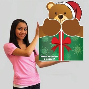 May include: A cartoon bear wearing a Santa hat peeks over a green gift box with a red bow. The box has the text "Want to know a secret?" The bear is brown with a white and red hat. The gift box has a green background with white snowflakes.
