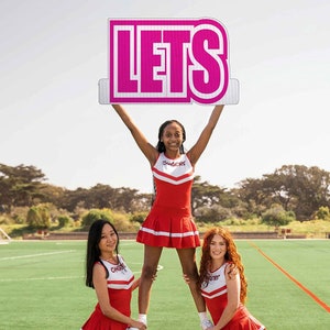 May include: Three cheerleaders in red and white uniforms are holding up a pink sign that says "LETS". The cheerleaders are on a green field with trees in the background.