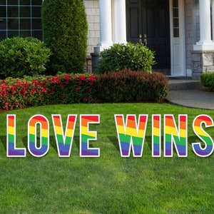 May include: A yard sign with the words "LOVE WINS" in large, rainbow-colored letters. The letters are white with a rainbow stripe pattern.