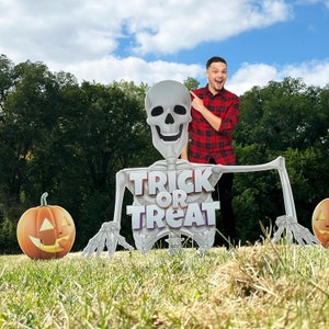 May include: A person dressed in a red and black plaid shirt stands in front of a large skeleton cutout holding a sign that says "Trick or Treat". The skeleton is wearing a white shirt and black pants. There are two orange pumpkins on the ground in front of the skeleton.