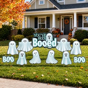 May include: A set of yard signs featuring white ghost figures with various facial expressions, including smiling and surprised faces, alongside two signs that say BOO and a central sign with the text YOU'VE BEEN BOOed in teal and white.