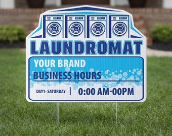 May include: A white and blue Laundromat sign with a lawn stake. The sign features illustrations of washing machines and the word "LAUNDROMAT" in large blue letters. The sign also includes space for business hours.