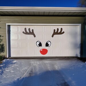 May include: A white garage door decorated with a reindeer face. The design includes brown antlers, black eyes with white highlights, and a large red nose. The driveway in front is covered in snow.
